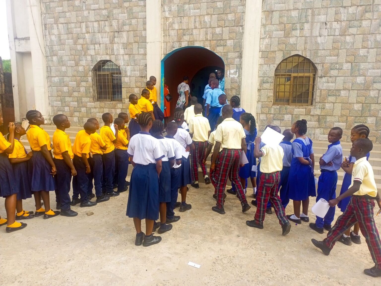 Zoe Robinson Academy students wearing pale yellow shirts and plaid pants walking toward the arched entrance of Jones Christian Academy to take their national examinations in Liberia.
